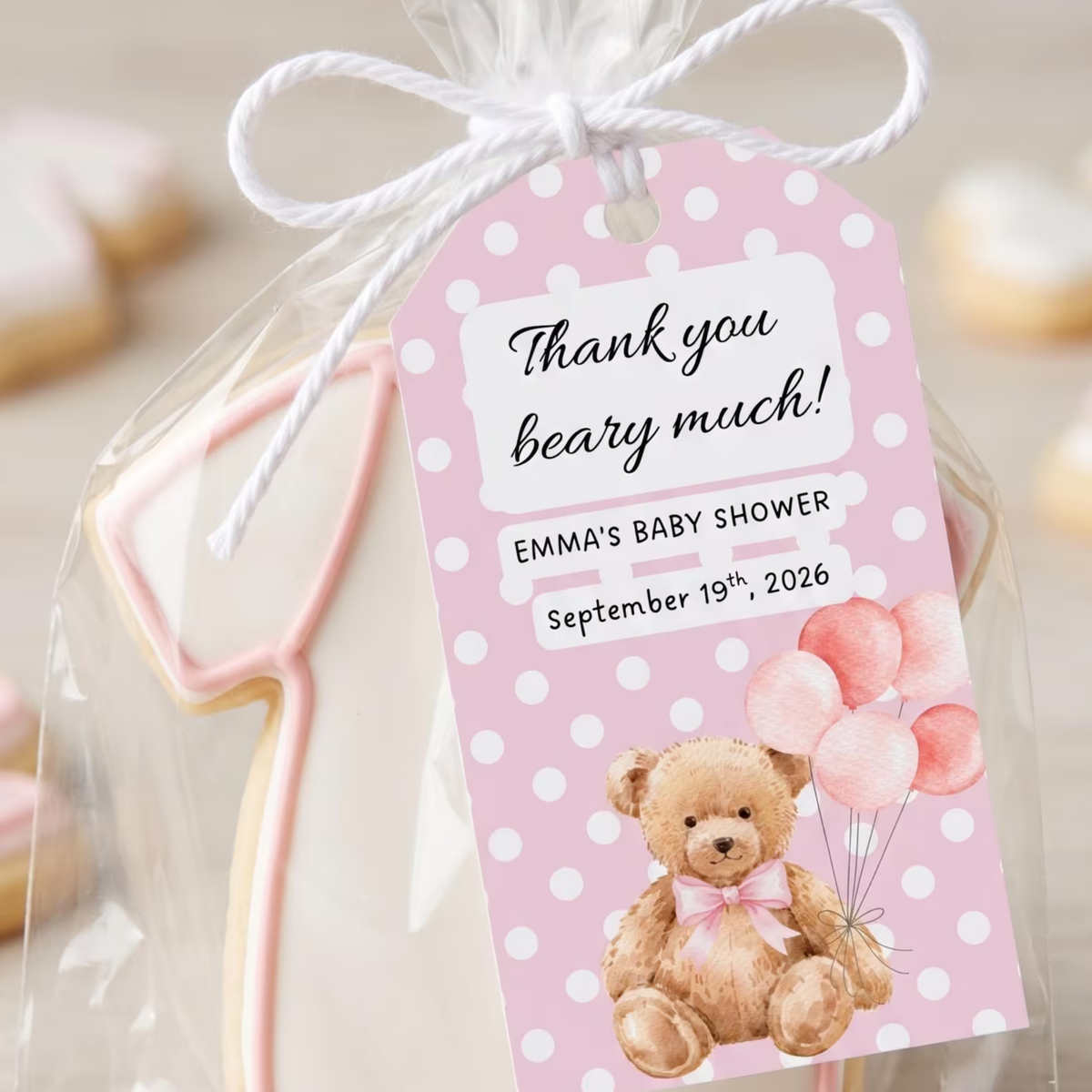 A personalized baby shower favor bag with a pink polka dot tag that reads 'Thank you beary much! Emma's Baby Shower September 19th, 2026' and features a teddy bear illustration. A pink and white cookie shaped like a stethoscope is visible inside the clear bag tied with a white string.