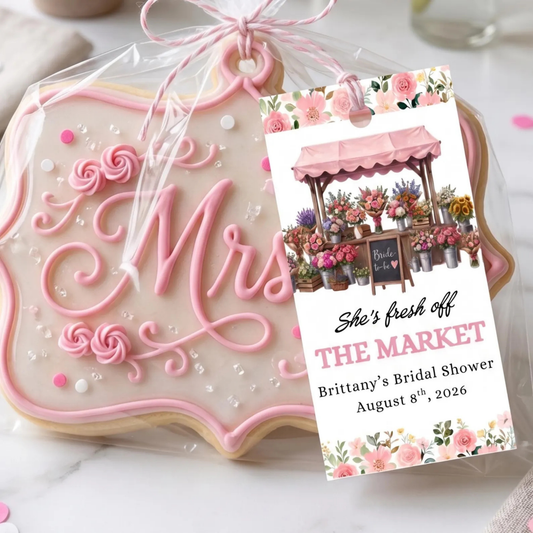 A decorated wedding cookie shaped like a bride's veil with pink icing and the word 'Mrs.' in script, wrapped in clear cellophane with a pink ribbon and a printed gift tag that reads 'She's fresh off THE MARKET Brittany's Bridal Shower August 8th, 2026'.