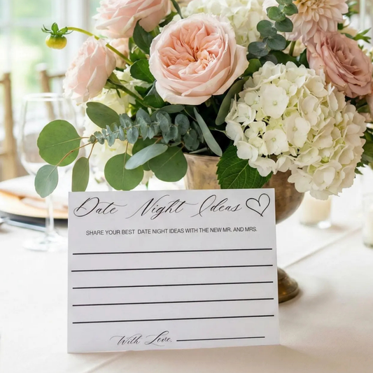 A white paper 'Date Night Ideas' table card with black script and lined writing space, placed in front of a floral centerpiece with blush pink roses, white hydrangeas, and green eucalyptus on a table setting.