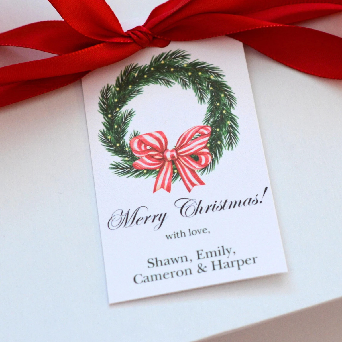 A personalized Christmas gift tag with a printed green wreath and red bow, tied with a red ribbon on a white gift box. The tag reads 'Merry Christmas! with love, Shawn, Emily, Cameron & Harper'.