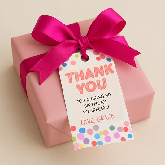 A pink gift box tied with a bright magenta satin ribbon and a white 'Thank You' tag printed with colorful polka dots, shown on a warm beige background.