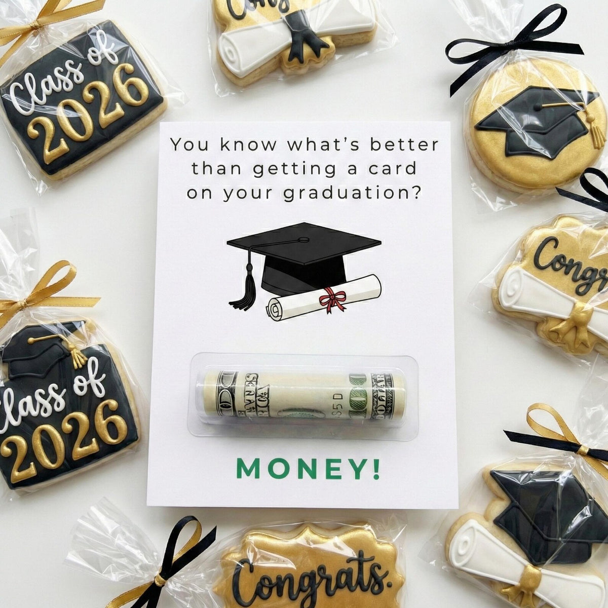 A graduation-themed cookie gift set featuring a white card that reads 'You know what's better than getting a card on your graduation? MONEY!' with a rolled dollar bill sealed in a clear plastic tube. The card is surrounded by decorated sugar cookies shaped like graduation caps, scrolls, and 'Class of 2026' and 'Congrats' cookies, each tied with black or gold ribbon bows.