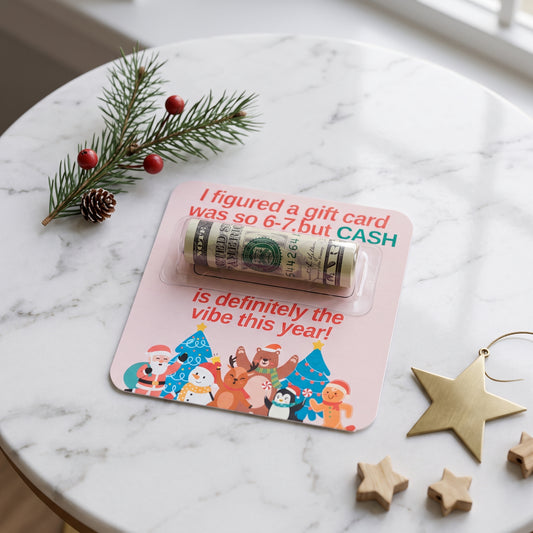 A rolled dollar bill sealed in a clear plastic money tube, mounted on a pink holiday gift card with festive animal illustrations and the text 'I figured a gift card was so 6-7,but CASH is definitely the vibe this year!'. The card is styled on a white marble table with pine branches, a pinecone, wooden stars, and a gold ornament.