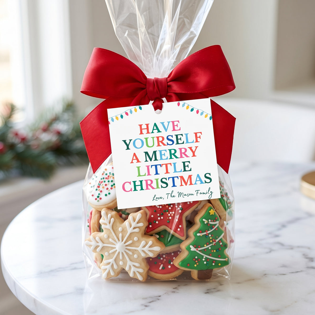 A clear cellophane gift bag filled with decorated Christmas sugar cookies, tied with a large red ribbon and a printed gift tag that reads 'HAVE YOURSELF A MERRY LITTLE CHRISTMAS' with the name 'Love, The Mason Family' below. The bag sits on a white marble table in a softly blurred indoor setting.