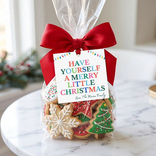 A clear cellophane gift bag filled with decorated Christmas sugar cookies, tied with a large red ribbon and a printed gift tag that reads 'HAVE YOURSELF A MERRY LITTLE CHRISTMAS' with the name 'Love, The Mason Family' below. The bag sits on a white marble table in a softly blurred indoor setting.