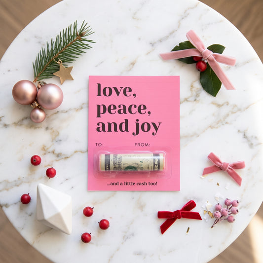 A pink holiday gift card with the words 'love, peace, and joy' and a rolled cash lip balm tube tucked inside, styled on a white marble surface with pink ribbons, greenery, ornaments, and small red berries.