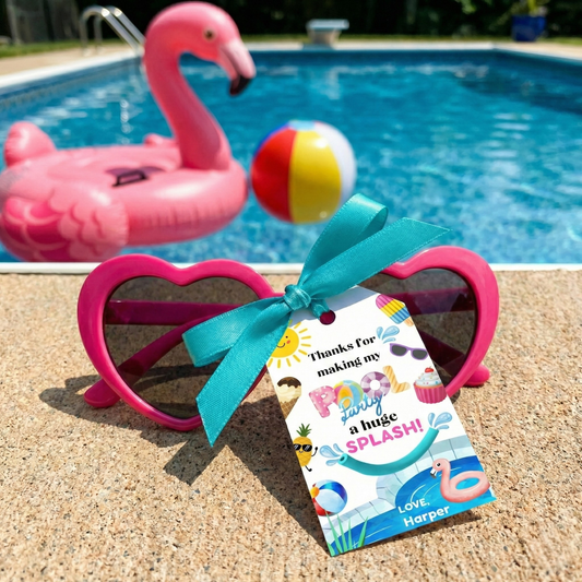 Pink heart-shaped sunglasses with a turquoise ribbon and a personalized gift tag, placed on a poolside deck with a pink flamingo float and beach ball in the background.
