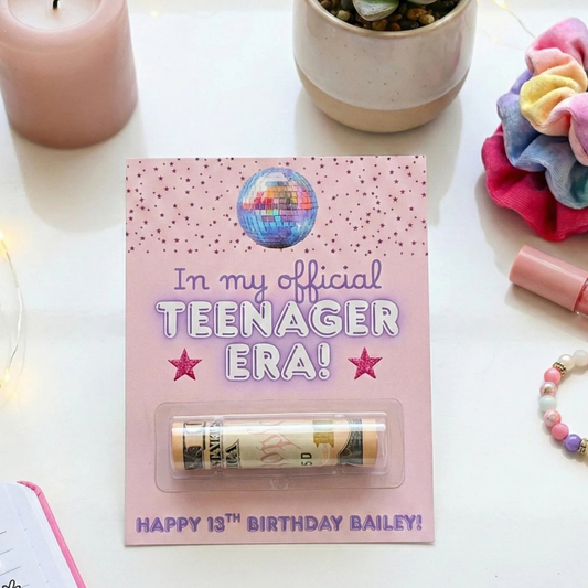 A pink personalized 13th birthday money tube gift card displayed on a white tabletop with decorative props. The card reads 'In my official TEENAGER ERA! HAPPY 13TH BIRTHDAY BAILEY!' with a disco ball graphic and a clear window showing a rolled dollar bill tube.