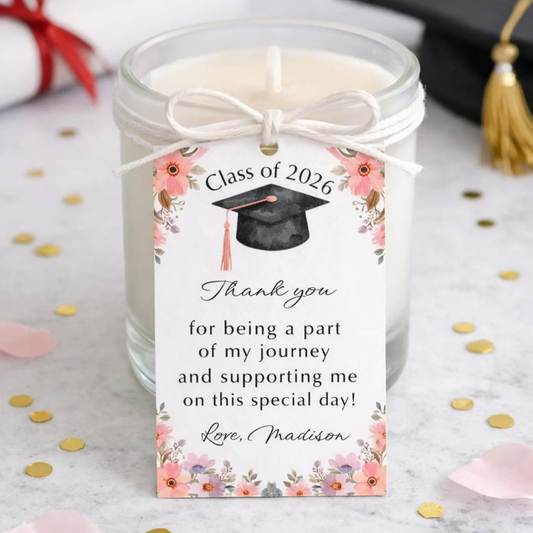 A glass jar candle with white wax and a white twine bow, featuring a printed graduation-themed tag that reads 'Class of 2026 Thank you for being a part of my journey and supporting me on this special day! Love, Madison' with floral illustrations. The candle is styled on a light marble surface with scattered gold confetti, pink petals, and blurred graduation accessories in the background.