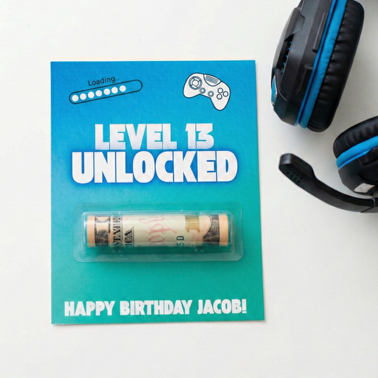 A custom video game themed birthday gift card featuring a 'Level 13 Unlocked' design with a rolled dollar bill inside a clear plastic capsule. The card is styled on a white surface next to a pair of black and blue gaming headphones.