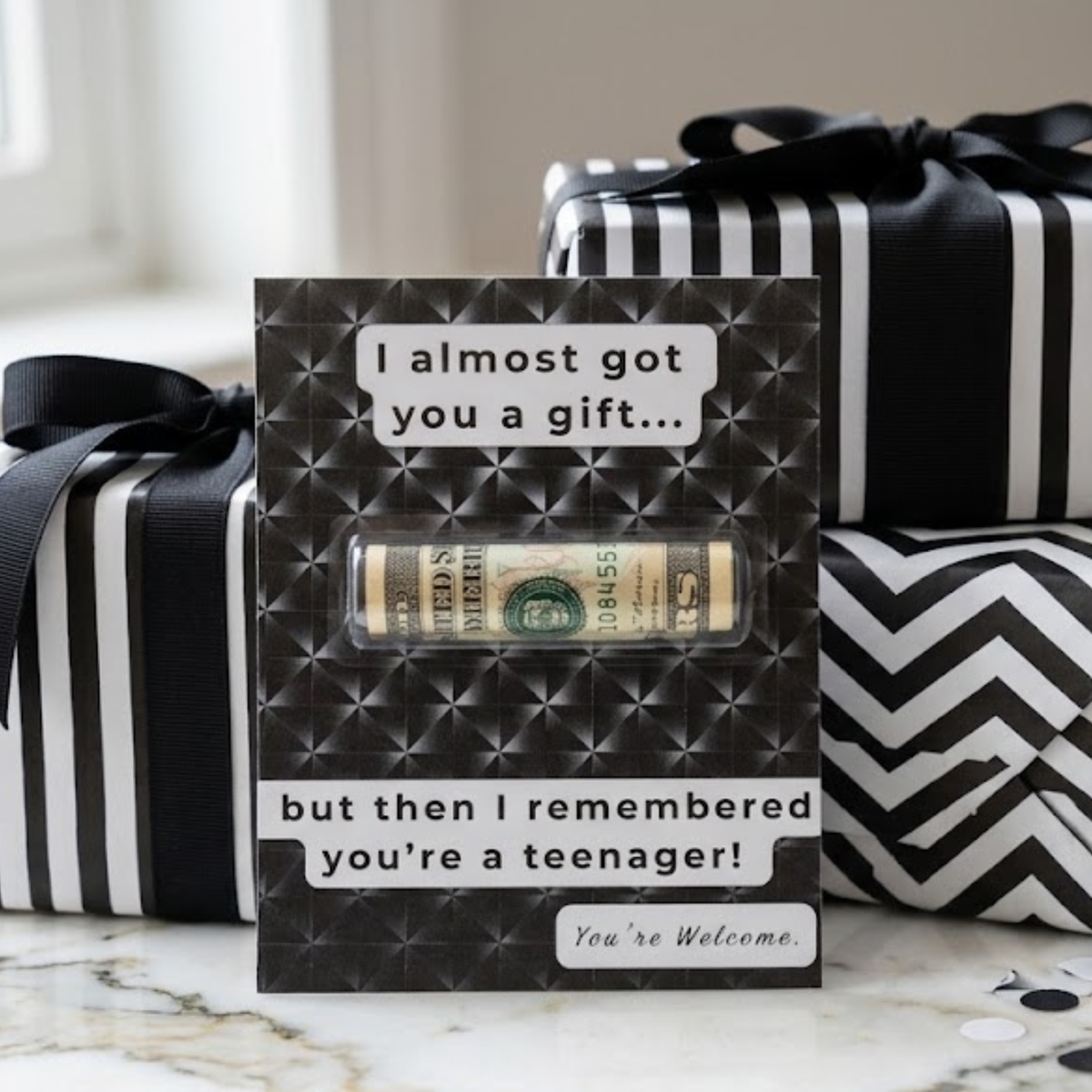 A novelty gift card with a rolled dollar bill window, styled in front of wrapped gift boxes with black and white patterns on a marble surface.