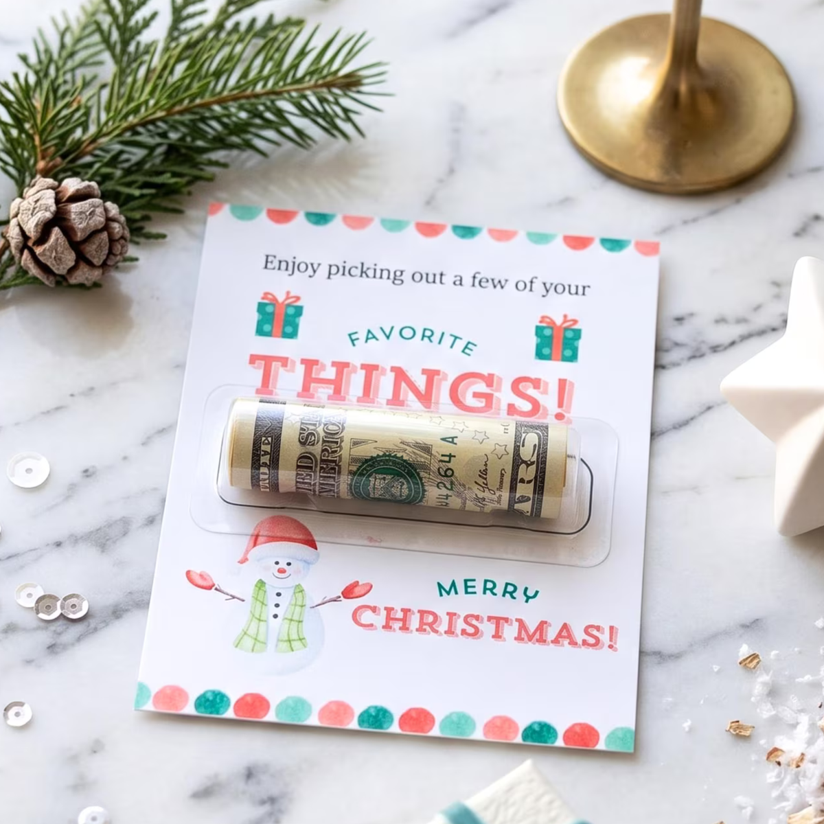 A rolled dollar bill in a lip balm tube attached to a festive Christmas-themed card that reads 'Enjoy picking out a few of your FAVORITE THINGS! Merry Christmas!'. The card is styled on a marble surface with pine branches, a pinecone, a gold candle holder base, and a white star ornament nearby.