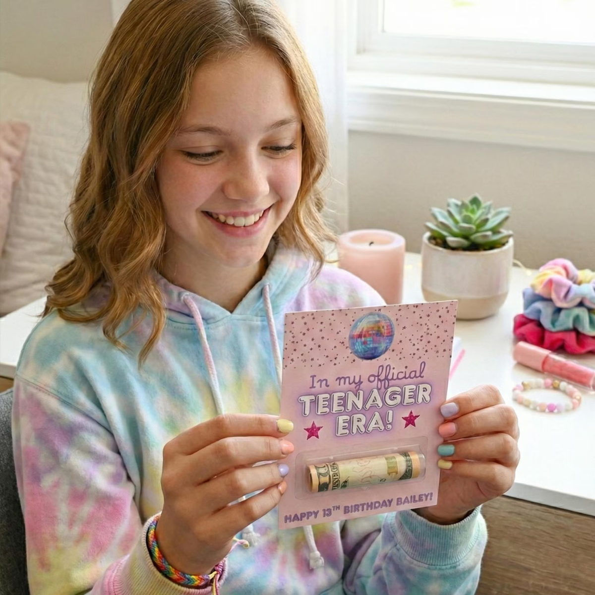 A smiling young woman holds a pink 'In my Official Teenager Era!' birthday card with a rolled dollar bill inside, in a bright indoor room with a window, candles, and small plants in the background.