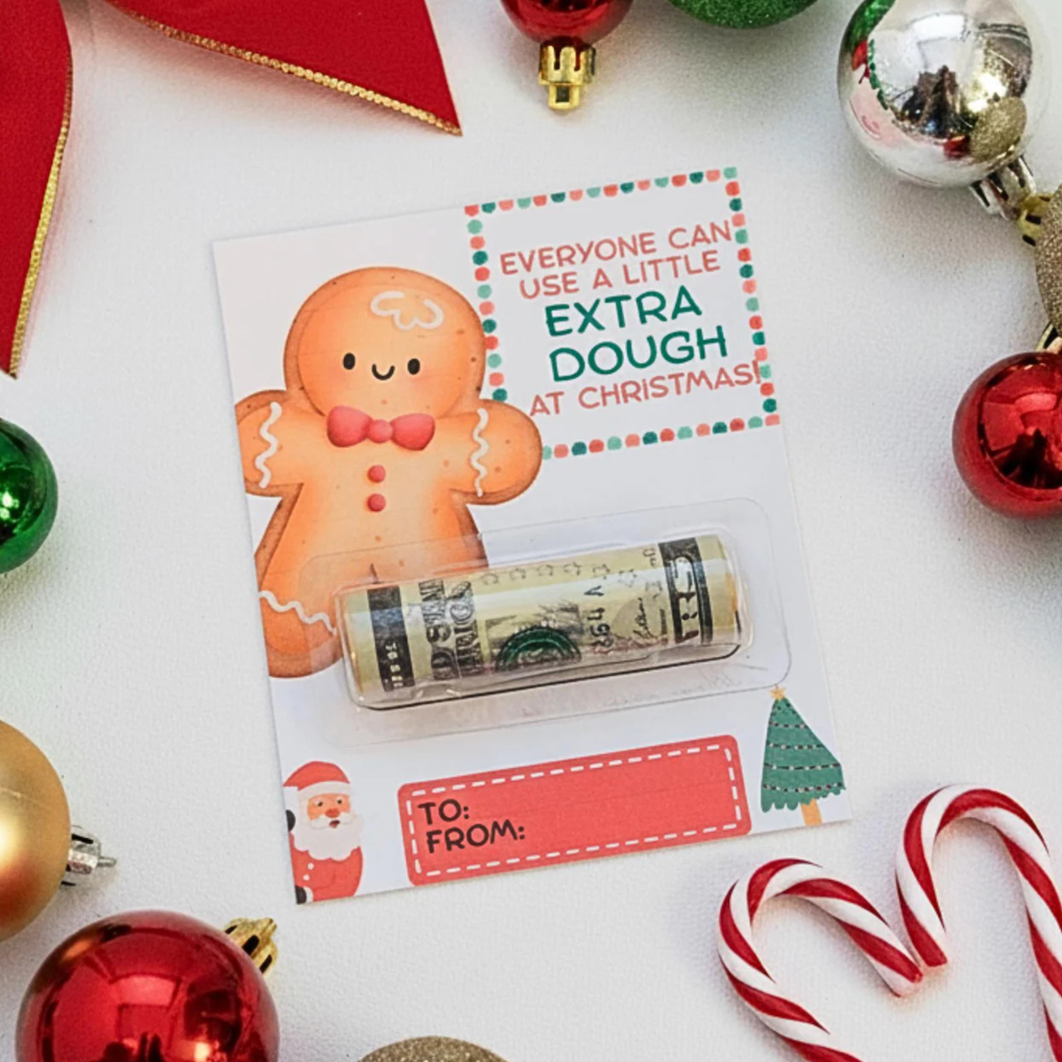 A Christmas-themed gift card featuring a gingerbread man illustration and the text 'Everyone can use a little extra dough at Christmas,' holding a rolled dollar bill under a clear plastic window. The card is surrounded by holiday decorations including red ornaments, candy canes, and a red ribbon on a white background.