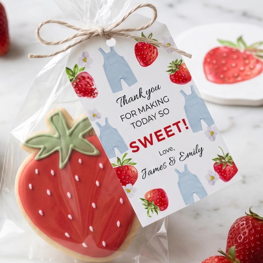 A strawberry-shaped sugar cookie in a clear bag with a twine-tied tag that reads 'Thank you FOR MAKING TODAY SO SWEET! Love, James & Emily.' The tag has strawberry and overalls illustrations, and the cookie is decorated with red icing and green leaf detailing on a light marble surface.