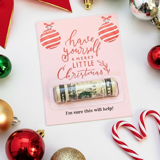 A rolled dollar bill money box sealed in a clear plastic window on a pink holiday card that reads 'have yourself A MERRY LITTLE Christmas' with red ornament illustrations. The card is photographed on a white surface surrounded by red, green, gold, and silver Christmas ornaments and candy canes.