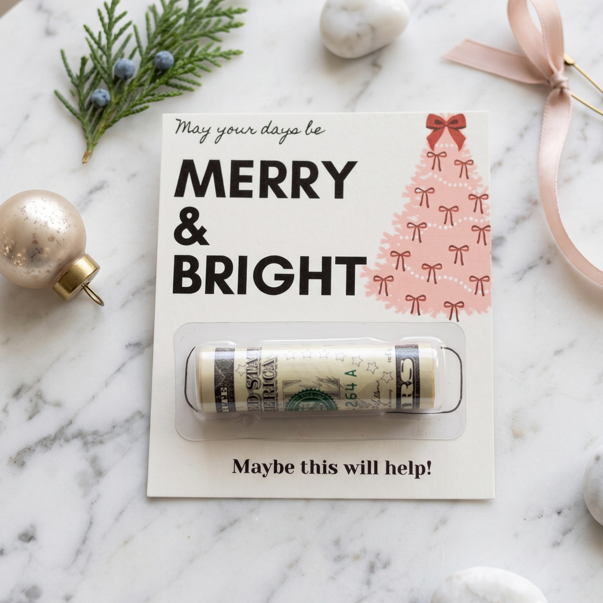 A rolled dollar bill money box wrapped in clear plastic, attached to a white holiday card that reads 'MERRY & BRIGHT' with a pink bow-tied tree illustration. The card is photographed on a white marble surface with Christmas ornaments, evergreen sprigs, and a pink ribbon around it.