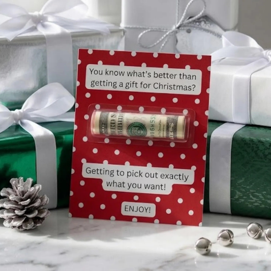 A red holiday gift card with white polka dots and text sits on a marble surface surrounded by wrapped presents, a pinecone, and silver bells. The card has a clear window showing a rolled dollar bill inside, with additional gift boxes in the background.