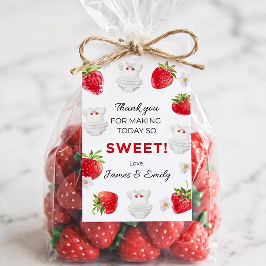 A clear cellophane bag filled with red strawberries and tied with a twine bow, featuring a printed thank-you tag with strawberries, wedding cake illustrations, and the text 'Thank you FOR MAKING TODAY SO SWEET! Love, James & Emily' on a light marble background.