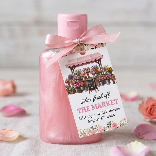 A pink bottle of bridal shower favor or gift set with a pink satin ribbon and a printed tag reading 'She's fresh off THE MARKET Brittany's Bridal Shower August 8th, 2026', styled with rose petals on a light surface.