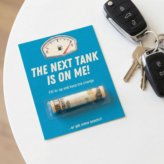 A blue novelty card on a white table next to car keys and keys in a purse, featuring the text 'THE NEXT TANK IS ON ME!' and a small rolled dollar bill sealed under a clear plastic cover.