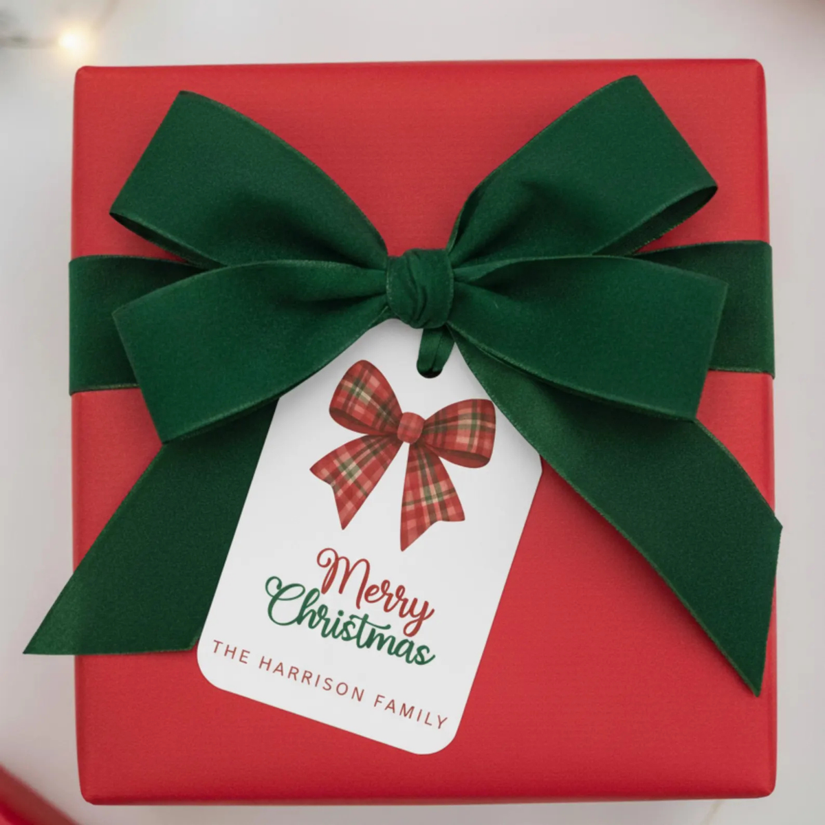 A red gift box wrapped with a large dark green ribbon and bow, featuring a white hanging tag that reads “Merry Christmas” and “THE HARRISON FAMILY,” photographed on a light background.