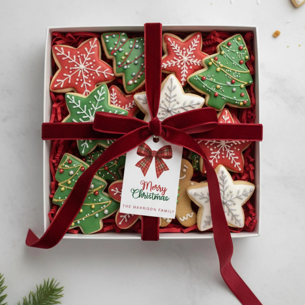 A gift box of decorated Christmas sugar cookies shaped like snowflakes and trees, wrapped with a red velvet ribbon and a hanging tag that reads 'Merry Christmas The Harrison Family,' on a light gray surface.