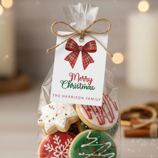 A clear cellophane gift bag filled with decorated holiday cookies and tied with a jute twine bow, featuring a white tag that reads 'Merry Christmas' and 'THE HARRISON FAMILY' with a red plaid bow illustration.