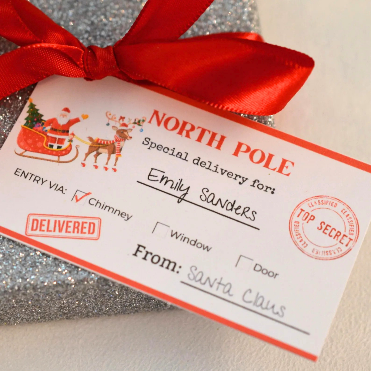 A close-up of a Christmas-themed gift tag reading 'NORTH POLE Special delivery for: Emily Sanders' and 'From: Santa Claus,' tied with a red ribbon over a silver glitter wrapped package.