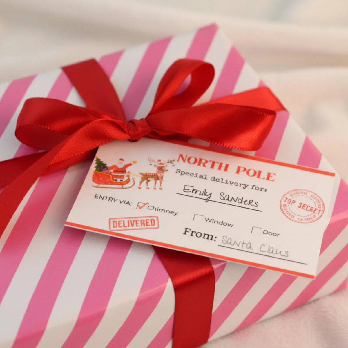 A wrapped gift box with pink and white diagonal stripes, tied with a large red ribbon and a novelty Santa-themed delivery tag that reads 'NORTH POLE' and 'Delivered from Santa Claus.'.
