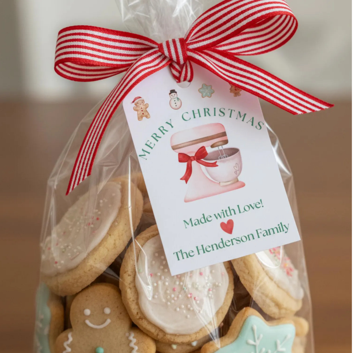 A clear cellophane gift bag filled with assorted decorated Christmas cookies, tied with a red-and-white striped ribbon and a printed tag that reads 'Merry Christmas Made with Love! The Henderson Family.'.