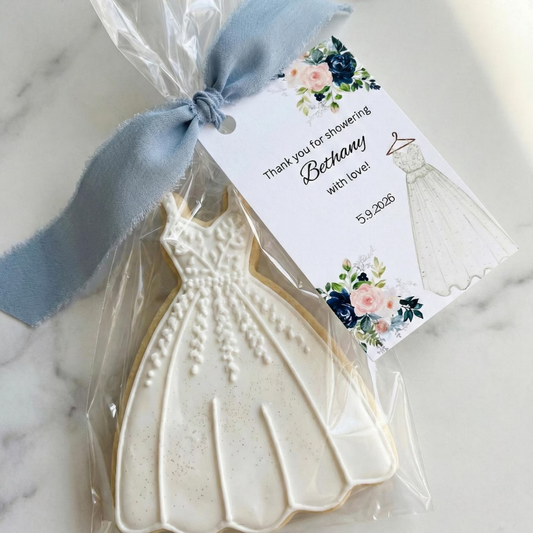 A shaped sugar cookie in the shape of a white wedding dress, wrapped in clear cellophane with a dusty blue ribbon and a printed thank-you tag, placed on a light marble surface.
