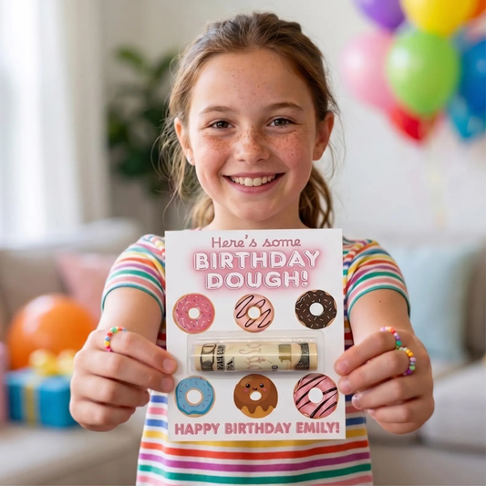 A smiling young girl with freckles holds up a novelty birthday card shaped like a doughnut wrapper. The card reads 'Here's some BIRTHDAY DOUGH!' and 'HAPPY BIRTHDAY EMILY!' with illustrated donuts and a rolled doughnut design.