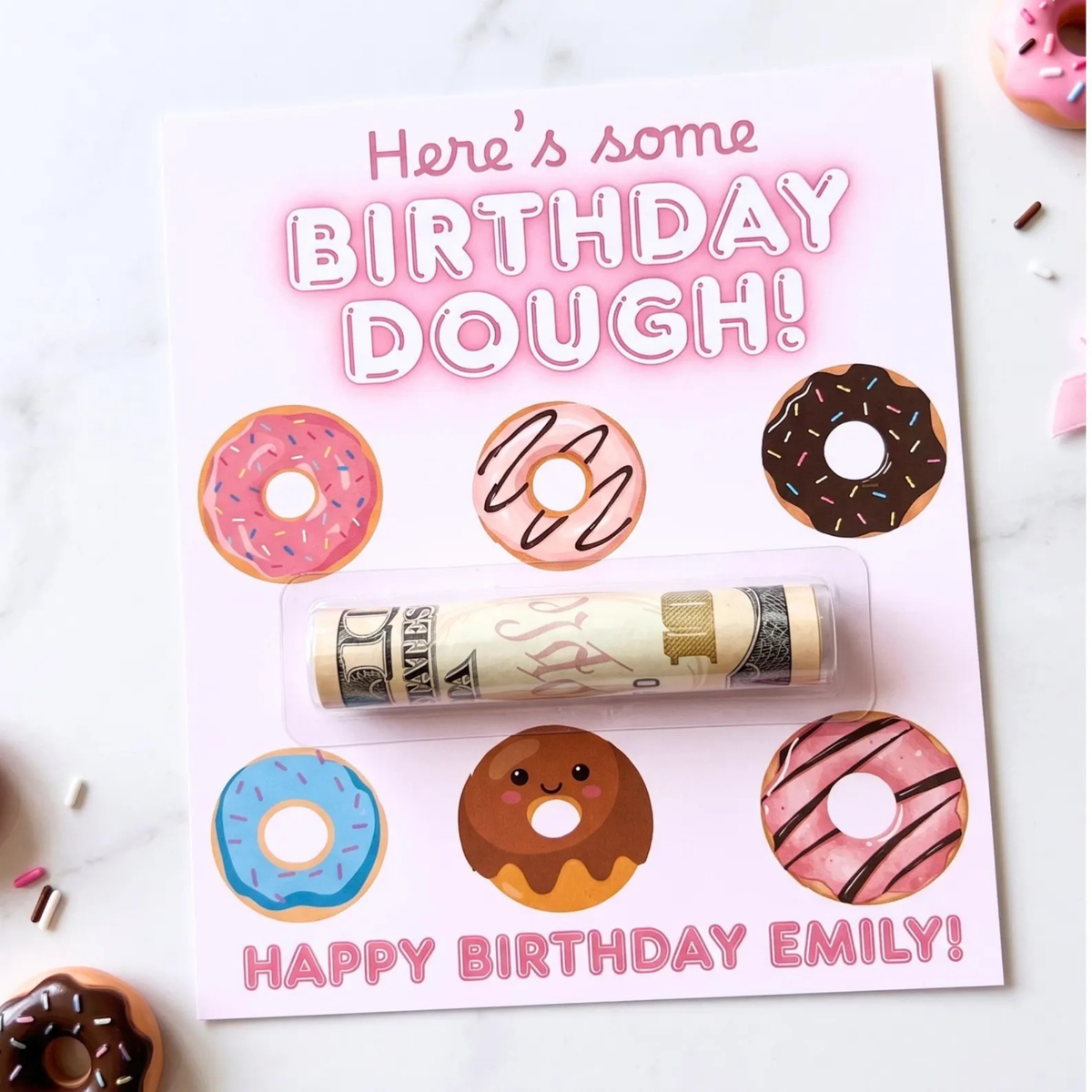A novelty birthday gift card featuring illustrated donuts and the text 'Here's some BIRTHDAY DOUGH!' and 'HAPPY BIRTHDAY EMILY!'. A rolled dollar bill is sealed in a clear plastic tube across the center.