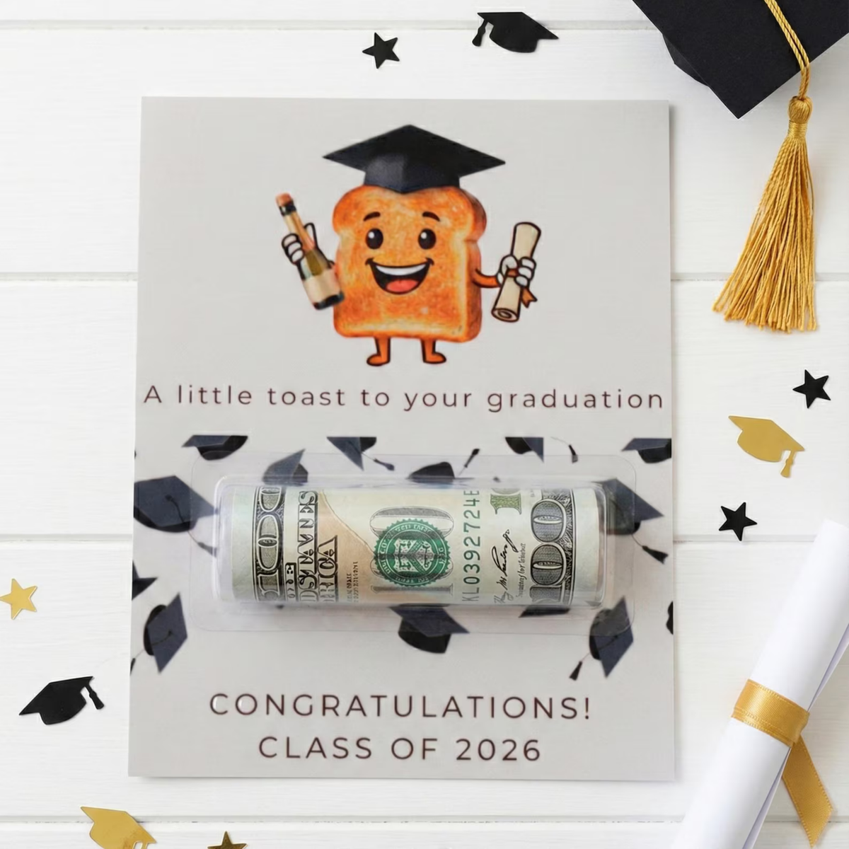 A graduation-themed card with a cartoon toast character wearing a graduation cap and holding a diploma and bottle. The card reads 'A little toast to your graduation' and 'CONGRATULATIONS! CLASS OF 2026,' with a rolled stack of cash tucked inside a clear plastic tube across the center.