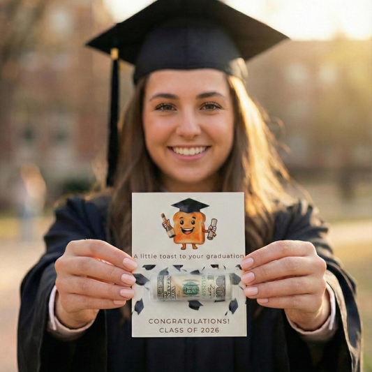 A smiling graduate in a black cap and gown holds up a custom cardboard money tunnel card featuring a cartoon graduation cap and the text 'A little toast to your graduation' and 'CONGRATULATIONS! CLASS OF 2026'.
