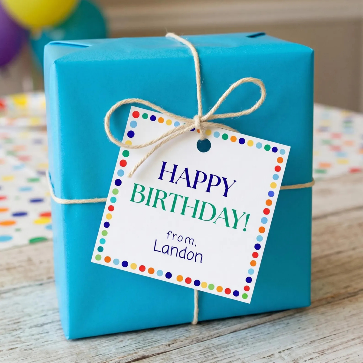 A bright turquoise gift box wrapped with natural twine and a white 'Happy Birthday!' tag with colorful polka dots, set on a wooden surface with a blurred party background.