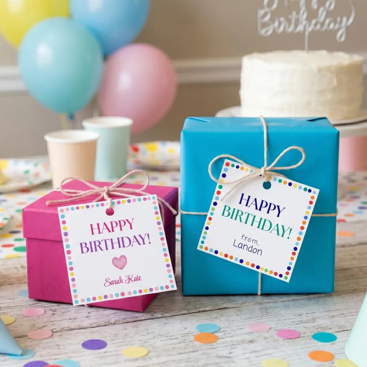 Two personalized birthday gift boxes on a party table, one magenta pink and one bright turquoise blue, both wrapped in paper and tied with twine, each featuring a colorful 'Happy Birthday!' gift tag. Balloons, cups, confetti, and a birthday cake are visible in the background.