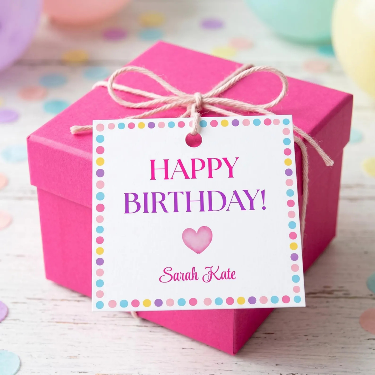 A bright pink gift box tied with twine and topped with a white 'HAPPY BIRTHDAY!' tag featuring a pink heart and the personalized name 'Sarah Kate', set on a light wood surface with pastel confetti and balloons in the background.