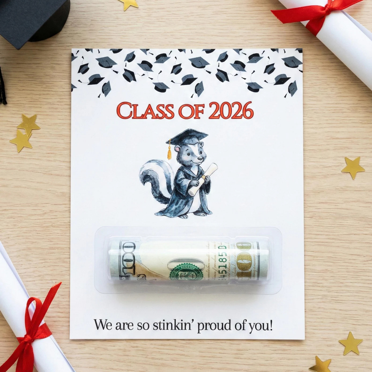A graduation-themed card or envelope on a light wood surface, featuring the text 'CLASS OF 2026,' a cartoon squirrel in a graduation cap, and a clear plastic window holding a rolled $100 bill. The card reads 'We are so stinkin' proud of you!' at the bottom.