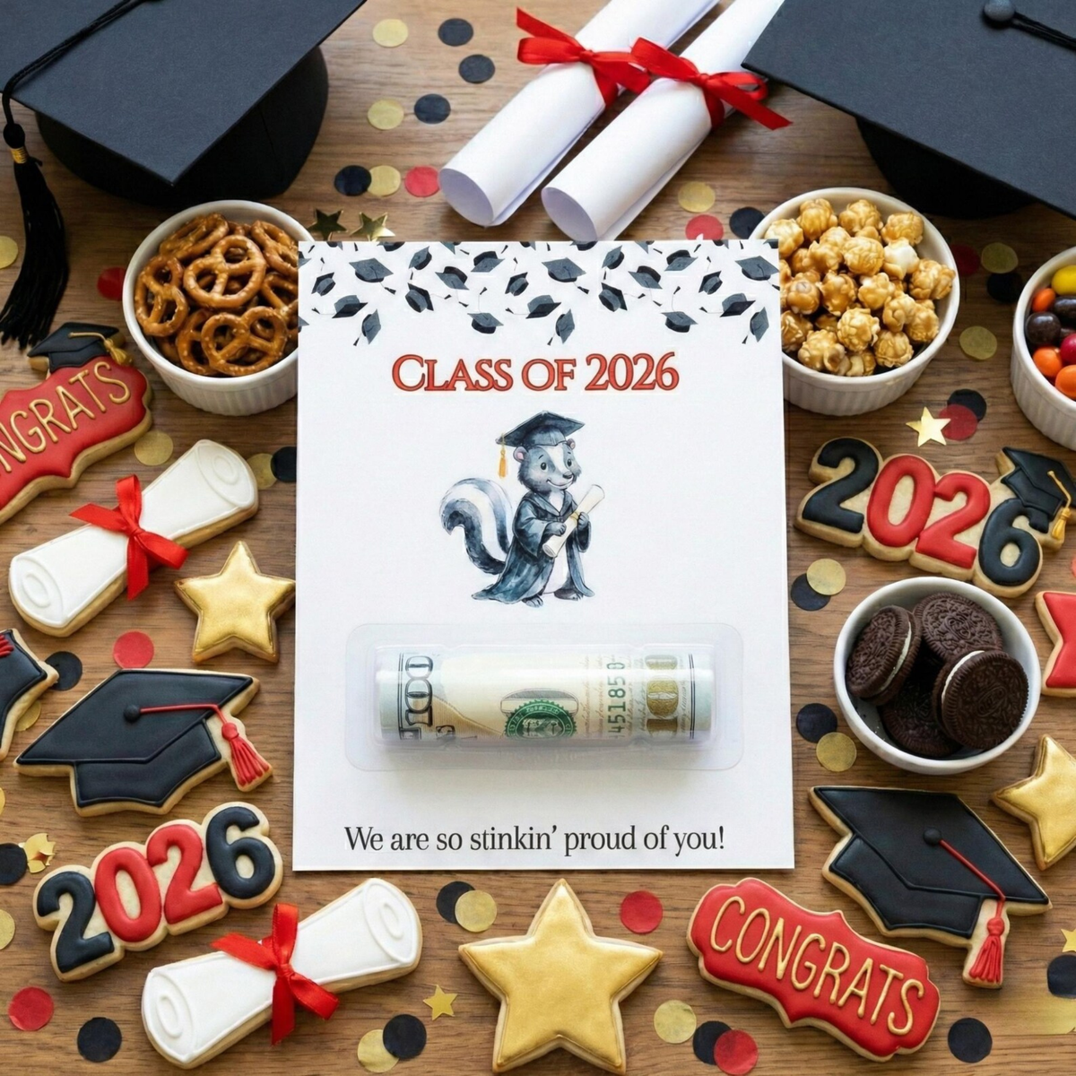 A rolled-up dollar bill sealed in clear plastic packaging, placed on a printed card that reads 'CLASS OF 2026' with a graduation cap illustration. The setup is a styled flat lay on a wooden surface surrounded by graduation-themed treats, cookie cutters, black mortarboard caps, rolled diploma tubes with red ribbons, pretzels, popcorn, Oreo cookies, candy, and decorative '2026' and 'CONGRATS' cookies.