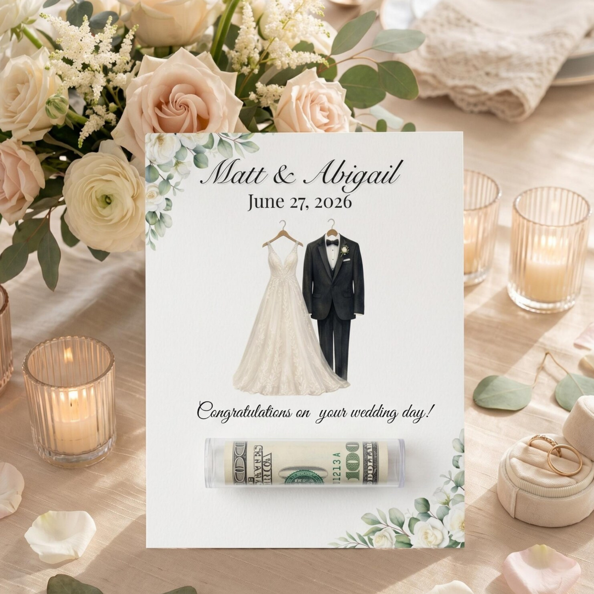 A personalized wedding keepsake card displayed on a wooden table surrounded by wedding flowers, candles, and rings. The card features the names 'Matt & Abigail' and the date 'June 27, 2020' above an illustration of a wedding dress and tuxedo, with the text 'Congratulations on your wedding day!' and a rolled dollar bill slot at the bottom.