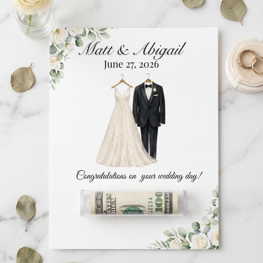 A personalized wedding money box on a white card, styled on a marble surface with decorative leaves, rings, and a vase. The card features a custom couple's wedding illustration with a bride and groom and the text 'Matt & Abigail June 27, 2026 Congratulations on your wedding day!'.