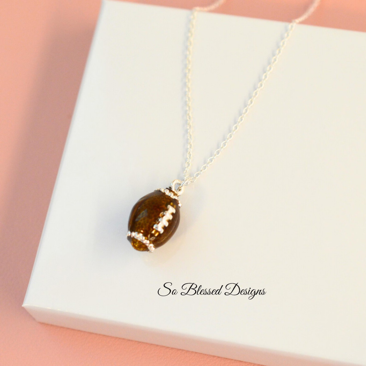 A silver chain necklace with an olive-shaped pendant featuring a brown center and white decorative stripes, displayed on a white gift box with the brand name 'So Blessed Designs' printed on it, set against a soft pink background.