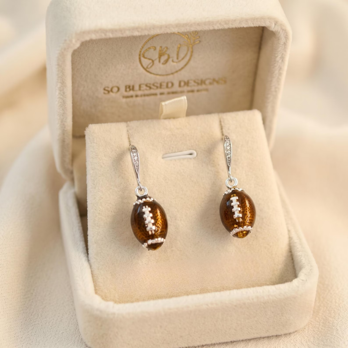 A pair of dangling football-shaped earrings with metallic amber-brown bodies and small silver-toned details, displayed in an open cream jewelry box. The inside of the box shows the So Blessed Designs logo.