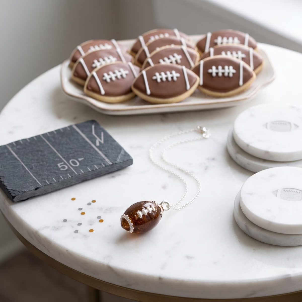 A football-shaped pendant necklace with a brown football design and white laces, displayed on a silver chain on a white marble table. In the background, a plate of football-shaped cookies sits on a tray, and a small slate yard-line prop lies nearby.