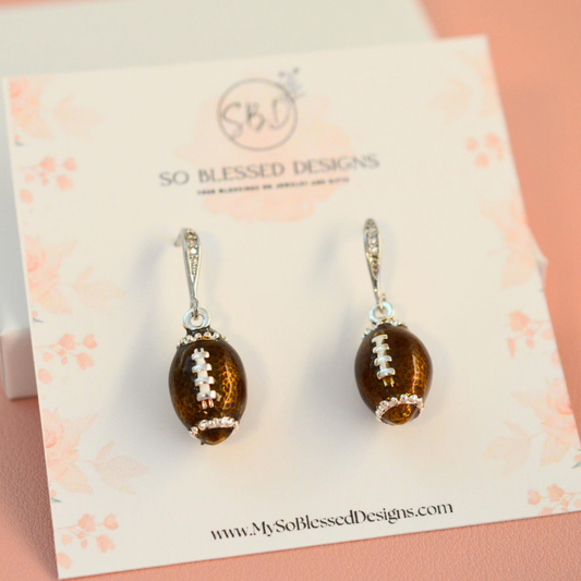 A pair of football-shaped drop earrings with amber-brown beads, silver-toned hooks, and white lacing details, displayed on a branded card from So Blessed Designs.