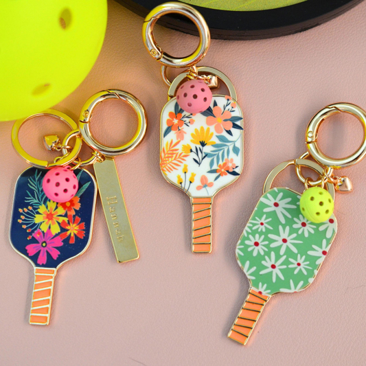 3 different pickleball keychains displayed near a pickleball racket and yellow pickleball. The first keychain is navy floral, the 2nd keychain is white floral with a light pink pickleball, and the 3rd keychain is green floral with a neon yellow pickleball.