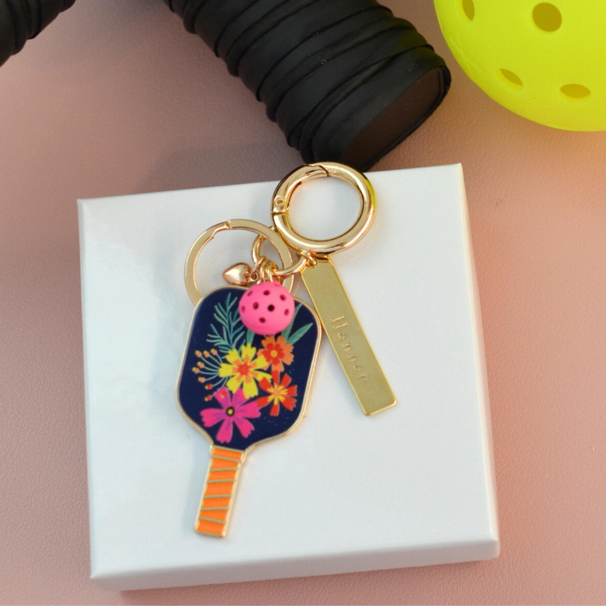 A navy floral pickleball keychain with a bright pink pickle ball with a gold name bar engraved with "Hannah" on it. All displayed on a white jewelry box. 
