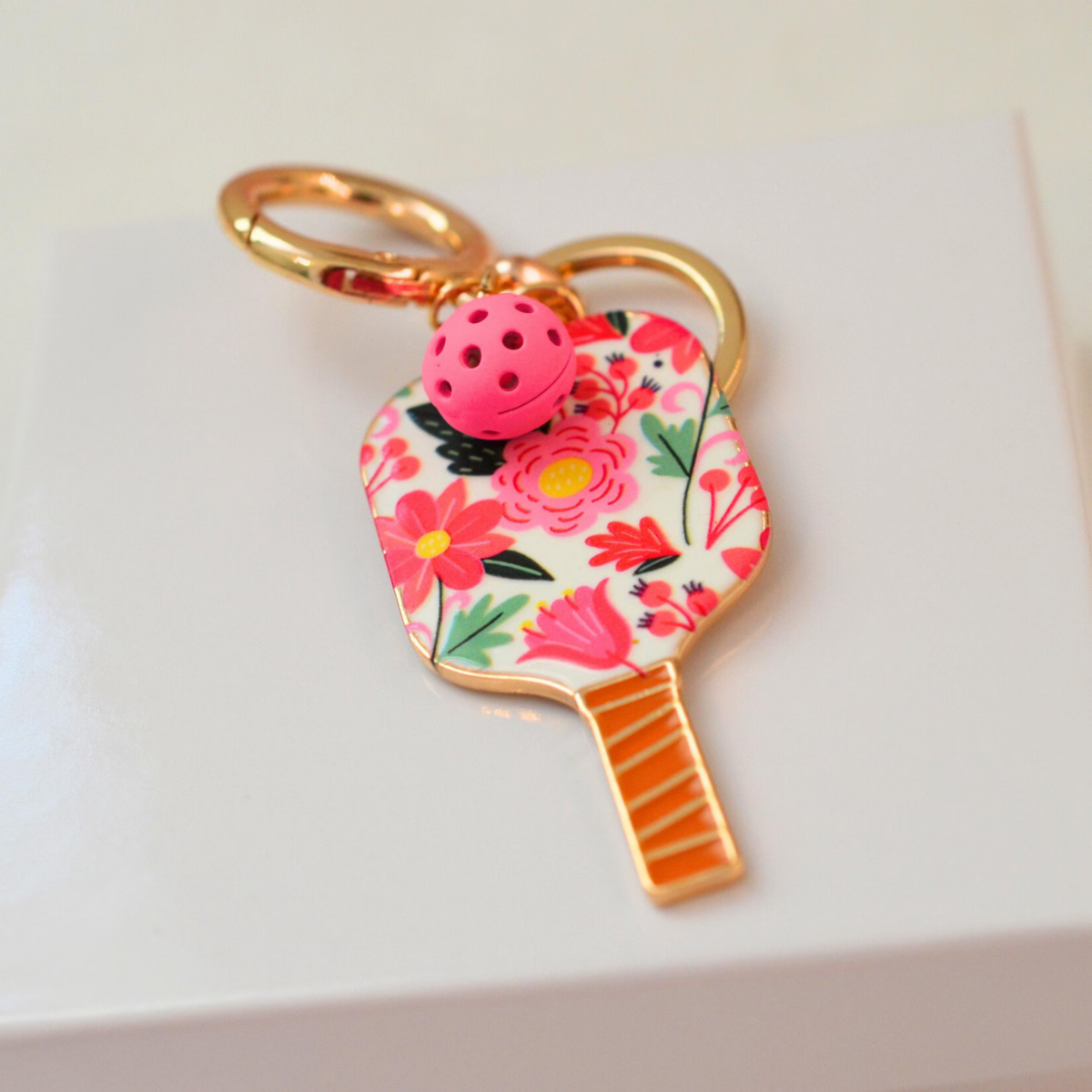 A tennis racket-shaped keychain charm with a pink and red floral pattern, gold-tone metal trim, a gold split ring, and a small pink perforated ball charm, resting on a white surface.